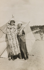 Kaksi naista väljissä, kirjavissa ranta-asuissa seisoo päivänvarjon edessä Hietarannassa